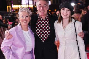 Dame Emma Thompson smiling at a film premiere alongside her husband, both dressed in formal attire, promoting her latest movie