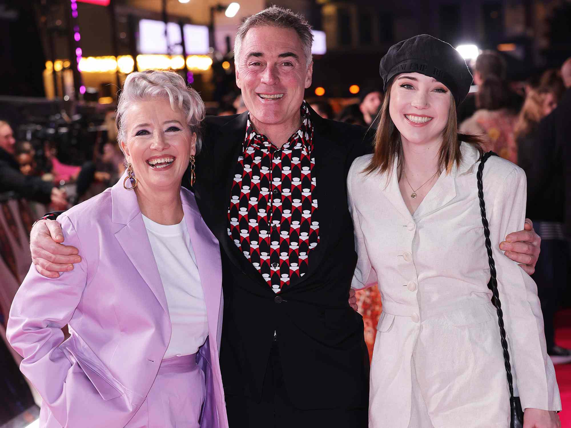 Dame Emma Thompson smiling at a film premiere alongside her husband, both dressed in formal attire, promoting her latest movie